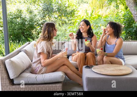 Diverse female friends laughing, chatting, holding drinks on rattan sofa on patio, phone on ottoman Stock Photo