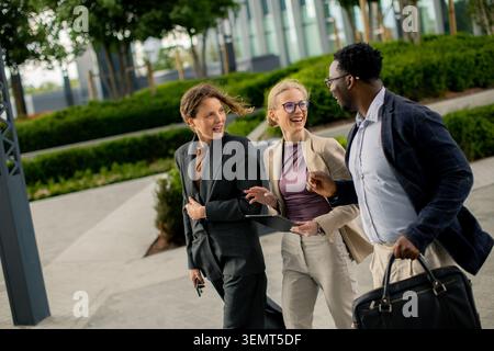 Three business people walk side by side in an urban park. They share smiles and laughter while holding briefcases and a tablet. Trees and modern build Stock Photo