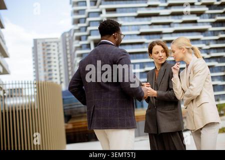 A group of three professionals stands outside modern buildings. They are engaged in discussion, sharing insights and ideas while looking focused. The Stock Photo