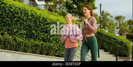 Two friends jog together on a pathway lined with greenery in a park during the evening hours. They appear to be having a good time while exercising ou Stock Photo