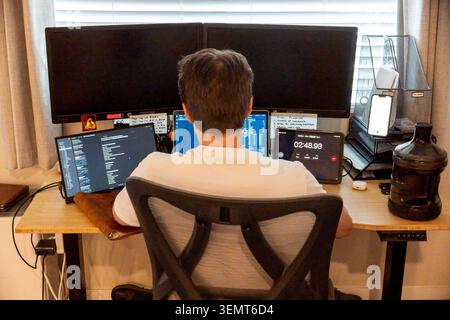 A person sits with back to camera at a cluttered desk featuring multiple computer monitors, a laptop, and office gear in a bright room Stock Photo