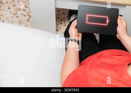 Person is holding a digital tablet displaying a low battery symbol while sitting on a sofa Stock Photo