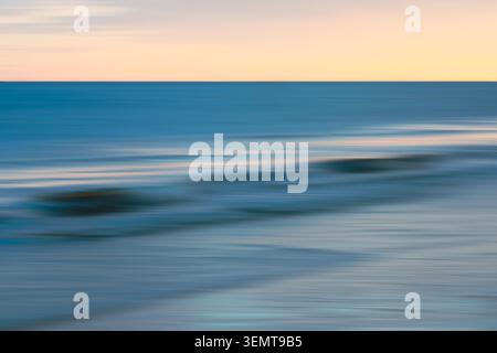 Blurred waves and shoreline reflect golden hour colors over the water. Stock Photo