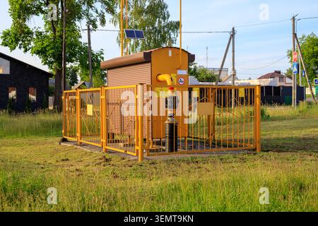 Yellow gas distribution box stands enclosed behind a safety metal fence. Rural pipeline regulator station on a village green grass lawn with houses an Stock Photo