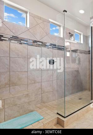 Bright bathroom showcases beige and gray tiled surfaces, a glass-walled shower stall, a built-in bench, and tall narrow windows that invite daylight. Stock Photo
