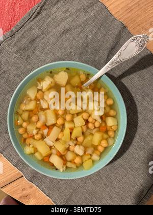 Bowl holds a warm potato and chickpea soup. The soup has chunks of potato and chickpeas in broth. A spoon rests in the bowl. Nearby is a glass filled with a drink Stock Photo