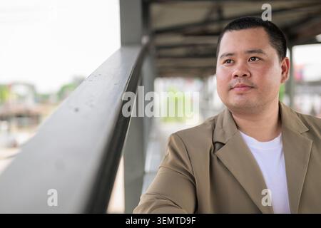 A confident plus size mid adult Asian businessman from the Isan region stands thoughtfully by a metallic railing on an overpass with a shallow depth o Stock Photo