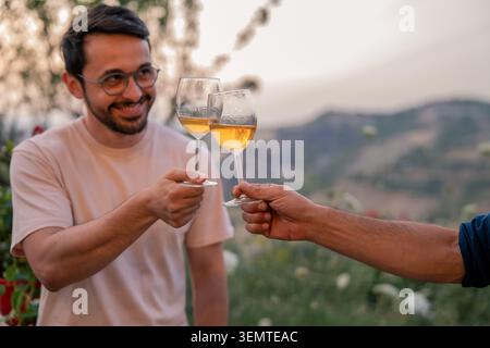 Two young adult men share a cheerful toast with glasses of white wine on an outdoor terrace in Sicily, Italy, with a scenic mountain landscape visible Stock Photo