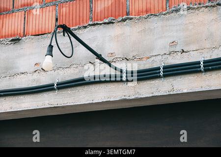 Exposed light bulb hanging with electrical wires and conduits on a concrete slab and a brick wall, showing industrial infrastructure Stock Photo
