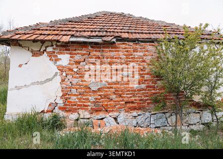 Derelict building standing in overgrown grass, featuring exposed red bricks, peeling plaster, and a damaged tiled roof Stock Photo