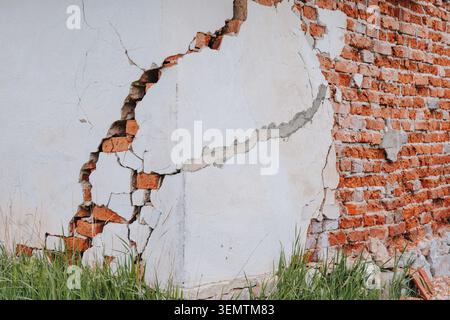 Peeling white plaster exposing a severely cracked, damaged red brick wall with green grass growing at its base, showing decay and neglect Stock Photo