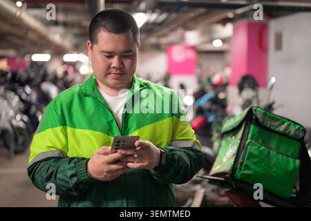 A focused plus size young adult Thai delivery man from the Isan region uses his smartphone next to a green insulated food delivery backpack inside a m Stock Photo