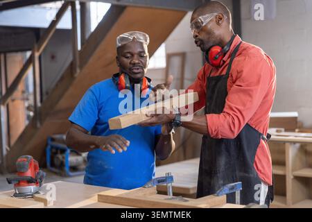 African American mentor and apprentice wearing goggles examining plank in workshop using sander Stock Photo