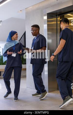 Diverse medical staff in navy scrubs discussing shift near elevator in lobby with tablet and badges Stock Photo