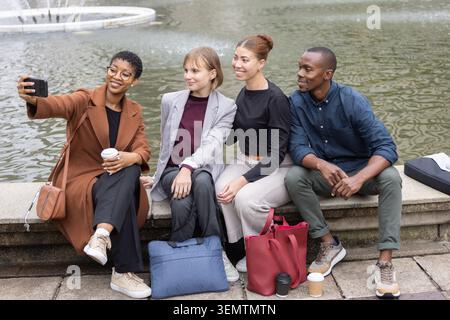 Diverse friends posing for selfie on stone ledge by fountain, holding smartphone and coffee Stock Photo