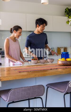 Diverse couple standing in home kitchen at island, in navy and pink, reaching bowls with berries Stock Photo