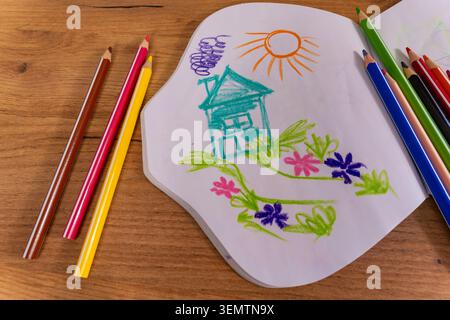 A colorful child's drawing of a house, flowers, and the sun in an open sketchbook surrounded by colored pencils on a wooden table. The cheerful artwor Stock Photo