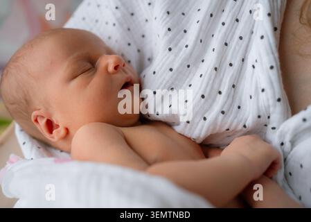 Newborn infant sleeping soundly wrapped in a white muslin cloth, nestled close to its mother's chest, symbolizing new life, maternal, and the tender bond between parent and child Stock Photo