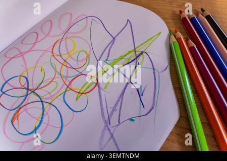 A close-up of colorful child scribbles in a sketchbook with colored pencils arranged on a wooden table. The abstract drawing captures early childhood Stock Photo
