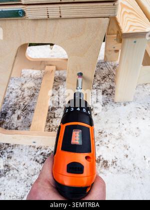 hand with electric screwdriver tightens screw while assembling chair outdoors close-up Stock Photo
