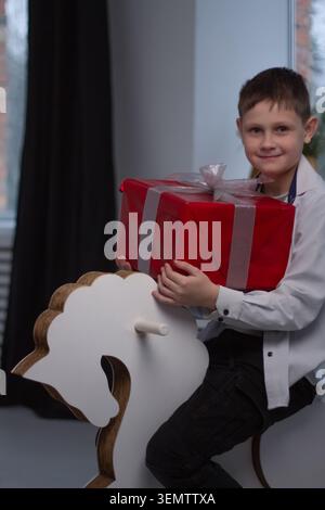 A boy sits on a white wooden rocking horse and holds a large gift in his hands. Stock Photo