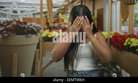 Young woman showing palms out in a stop gesture, hands near face among potted flowers and large planters in a building; fear hesitation. Stock Photo