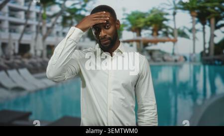 Man wiping forehead with hand at hotel building poolside in white shirt, face and beard visible, weary expression and slight fatigue; reflective pause Stock Photo