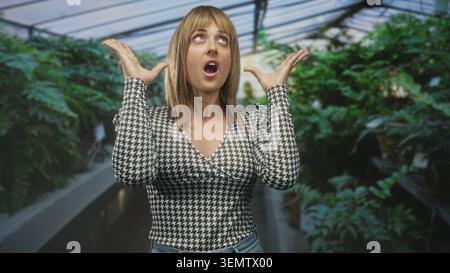 Woman with raised hands and open mouth looking up in a plant filled building aisle, wearing houndstooth top and jeans; wonder discovery nature. Stock Photo