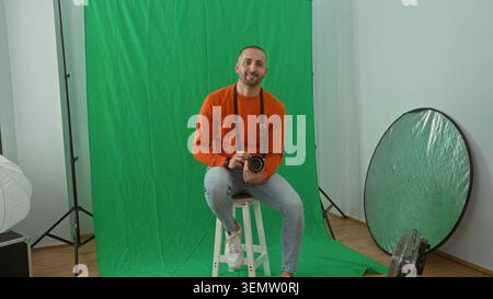 Man holding camera sits on stool in studio with green screen and reflector, smiling and leaning slightly forward; friendly relaxed casual. Stock Photo