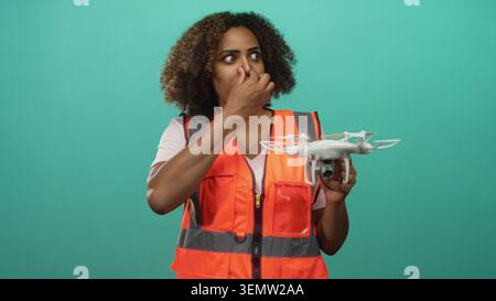 Young african american woman engineer holding a drone pinching nose in studio with teal backdrop and orange safety vest; disgust. Stock Photo