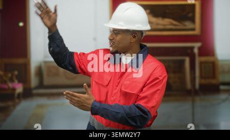 Man construction worker in red uniform and white hardhat gesturing with hands and pointing while presenting artwork in a museum gallery; instruction c Stock Photo