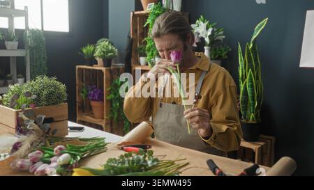 Man sniffing a pink tulip with nose and right hand, rubbing his eye and wincing while holding the stem in a flower studio; discomfort. Stock Photo
