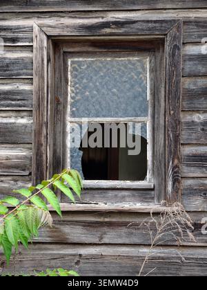 A single-pane window set in a worn, gray wooden wall with peeling boards and a cracked lower glass pane. The rustic frame shows aged grain and faded p Stock Photo