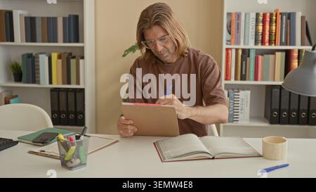 Young blond man student sitting at desk with open book, clipboard, pen and glasses, hand pressed to forehead in a building; frustration. Stock Photo