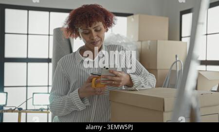Woman in striped shirt holds smartphone in right hand and yellow mug in left while leaning on moving boxes in building looking at screen; moving day c Stock Photo