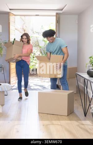 Couple carrying cardboard moving boxes through open front door into foyer, wearing casual clothing. Entryway, sunlit, unpacking, cartons, home, minima Stock Photo