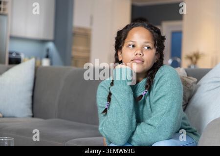 Female child sitting on gray sectional sofa in living room, wearing teal sweater, purple hair ties Stock Photo