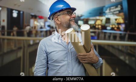 Man architect holding rolled blueprints and smiling with hand on plans in building, wearing blue hardhat; confidence planning future projects. Stock Photo