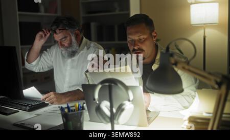Two men at a desk, one reads a document while the other rubs forehead beside a laptop in an office building; late night fatigue. Stock Photo