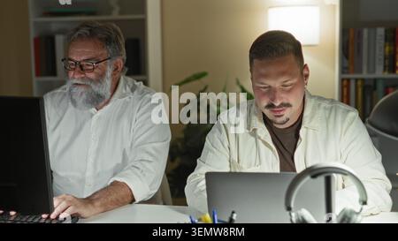 Two men at a desk working on laptop and desktop, older man typing while the younger studies the screen in a building; late night focus collaboration. Stock Photo