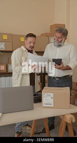 Two men examine shipping papers and a tablet amid stacked boxes on shelves, discussing orders and labels in a building storage room; teamwork focused. Stock Photo