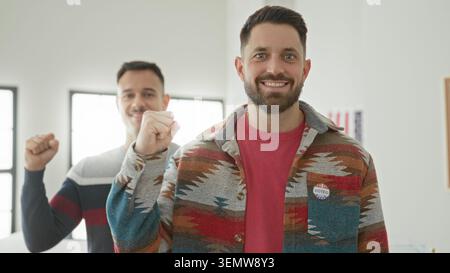 Two men showing i voted sticker and raised fists, one hand clenched near chest in polling building; civic pride. Stock Photo