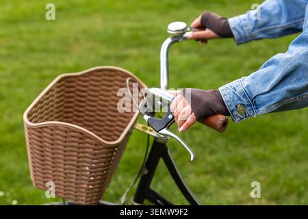 Hands Holding Vintage Bicycle Handlebars. Close-up of hands on vintage bicycle handlebars with a wicker basket, set against soft green grass outdoors. Stock Photo