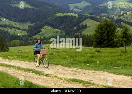 Woman Cycling Through Green Mountain Meadow. Smiling young woman rides a vintage bicycle along a dirt path through a scenic green mountain countryside Stock Photo