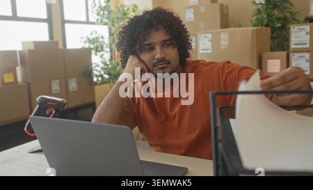 Man in orange shirt with laptop and cardboard box, hand on cheek while sorting paperwork in building; tired small business. Stock Photo