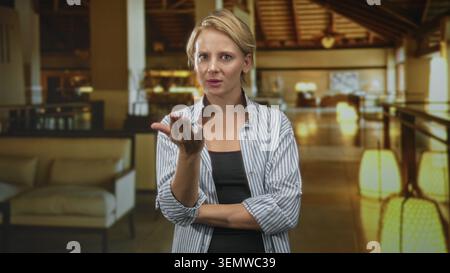 Woman extends hand palm up and beckons in a building lobby, arm crossed at waist; demanding expectation. Stock Photo
