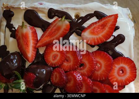Close-up top view of a delicious cake decorated with fresh sliced strawberries and melted dark chocolate topping on a light textured background. Stock Photo