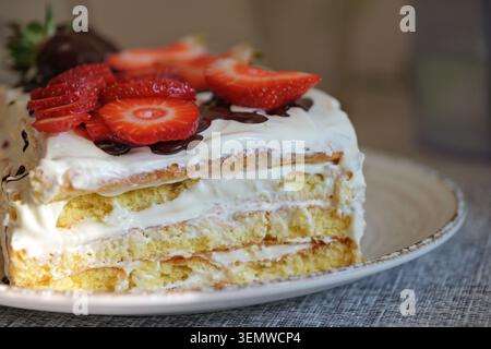 Close-up of a homemade layered sponge cake with creamy filling, fresh strawberry slices and chocolate drizzle. Appetizing dessert served on a plate wi Stock Photo
