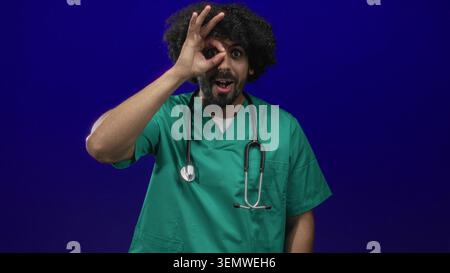 Man doctor in green scrubs with stethoscope making ok sign with hand over eye in studio with deep blue background; playful curiosity. Stock Photo