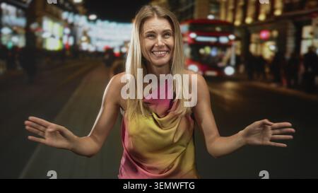 Blonde woman smiling and dancing with raised hands near a red double decker bus in a night city street; joyful. Stock Photo